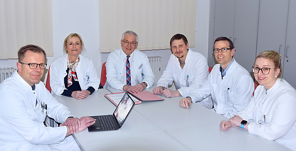 Sechs Ärzte im Kittel sitzen an einem Besprechungstisch und blicken in die Kamera